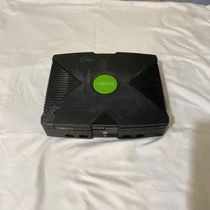 May include: Black Xbox console with a green circular logo in the center. The console has a rectangular shape with a ridged top and the word "XBOX" printed on the front. The console is sitting on a white surface.
