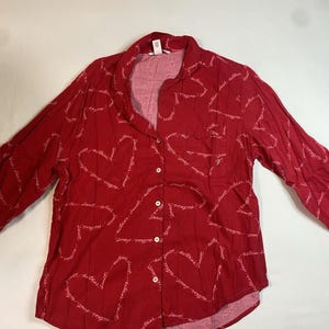 May include: A long-sleeved red button-down pajama shirt featuring a repeating white script pattern that forms heart shapes. The shirt includes a notched collar, a single chest pocket on the left side, and white buttons down the front center.