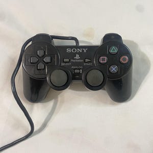 May include: A black wired Sony PlayStation game controller featuring a directional pad, two analogue sticks, four action buttons with triangle, circle, square, and cross symbols, and centre buttons labelled Select, Start, and Analog.