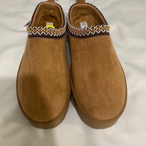 May include: A pair of tan slip-on shoes with a soft, textured exterior and a thick, matching tan sole. The collar of each shoe features a decorative brown trim with a white and gold cross-stitch pattern. The interior is lined with tan faux fur.