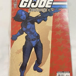 May include: A comic book cover for G.I. Joe: Origins, featuring a blue-toned female character holding a weapon. The cover has a red and gold star-patterned background. The text includes "HAMA * HAWTHORNE" and "GI JOE ORIGINS".