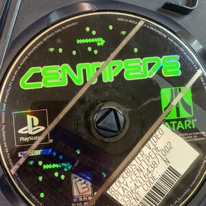Peut inclure: Un disque de jeu PlayStation noir pour Centipede, avec du texte vert et blanc. Le disque est dans un boîtier noir avec un porte-carte mémoire. Les logos Atari et PlayStation sont visibles.