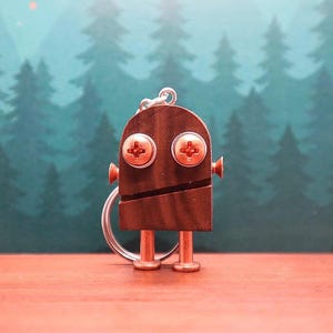 May include: A handmade robot keychain made of brown wood and metal hardware. The robot has two large screw-head eyes, small ear-like screws, and two metal legs. The keychain is attached to a silver metal ring. The background is a blurred forest scene.