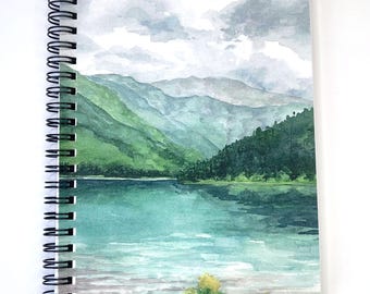 May include: Spiral-bound notebook featuring a watercolor painting of a mountain lake scene. The cover art depicts green mountains, a turquoise lake, and a cloudy sky. Yellow flowers are at the bottom. The notebook has a black spiral binding.