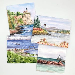 May include: Four watercolor paintings depicting coastal scenes. One shows a lighthouse on a cliff, another a lighthouse near a forest, a third a harbor with a lift bridge, and the fourth a lighthouse at the end of a stone pier by the water.