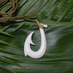 May include: A white, curved hook pendant necklace with a woven brown cord. The pendant is made of a smooth, glossy material. The necklace rests on a green leaf background, creating a natural aesthetic. The hook design suggests a nautical or island theme.