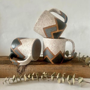 May include: Three ceramic mugs with a white speckled glaze and geometric patterns in black, brown, and gray. The mugs are stacked on top of each other on a wooden surface.