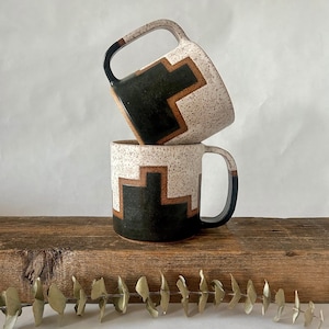 May include: Two ceramic mugs stacked on top of each other. The mugs are white with black and brown geometric designs.