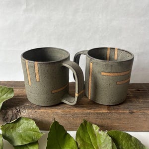 May include: Two handmade ceramic mugs. The mugs are a muted green color with tan-colored stripes. The mugs have a rounded shape with a handle on the side. The mugs are sitting on a wooden surface.