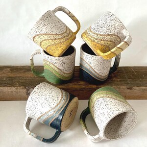 May include: Five ceramic mugs with speckled white glaze and colorful wavy designs. The mugs are stacked and arranged on a wooden surface. The designs include yellow, green, and blue.