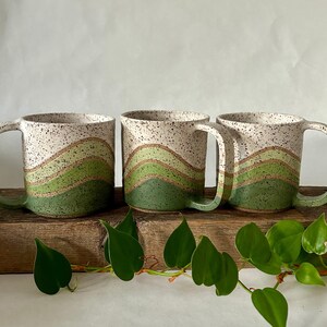 May include: Three white ceramic mugs with green and brown wavy designs. The mugs are sitting on a wooden surface with green leaves.