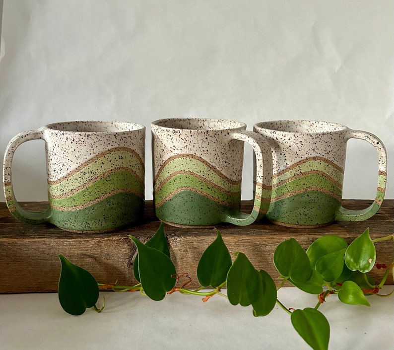 May include: Three white ceramic mugs with green and brown wavy designs. The mugs are sitting on a wooden surface with green leaves.
