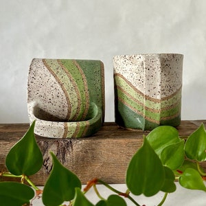 May include: Two ceramic mugs with a speckled white glaze and green and brown stripes. The mugs are sitting on a wooden surface.