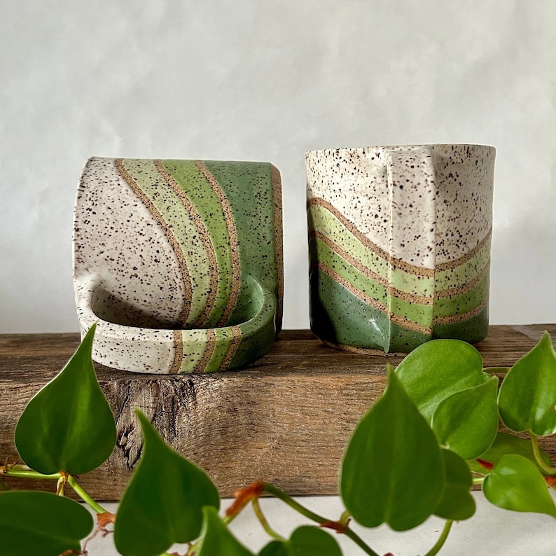 May include: Two ceramic mugs with a speckled white glaze and green and brown stripes. The mugs are sitting on a wooden surface.