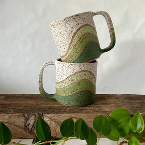 May include: Two white ceramic mugs with green and brown wavy designs stacked on top of each other. The mugs have speckled white glaze and are sitting on a wooden surface.