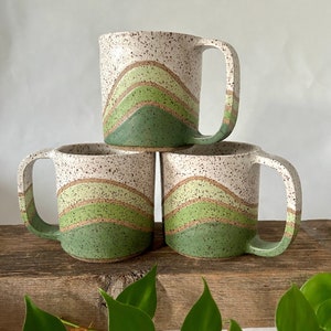 May include: Three ceramic mugs with a white speckled glaze and green and brown stripes. The mugs have a rounded shape and a handle.