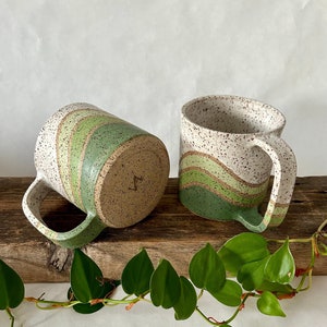 May include: Two ceramic mugs with green and white speckled glaze. The mugs have a wavy green stripe design. One mug is shown from the side and the other is shown from the bottom.