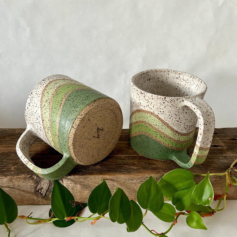 May include: Two ceramic mugs with green and white speckled glaze. The mugs have a wavy green stripe design. One mug is shown from the side and the other is shown from the bottom.