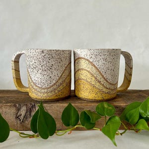 May include: Two white ceramic mugs with speckled brown glaze and yellow and brown wavy lines. The mugs have a handle on the right side.