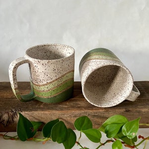 May include: Two ceramic mugs with a speckled white glaze and green and brown stripes. The mugs are sitting on a wooden surface with green plants in the foreground.