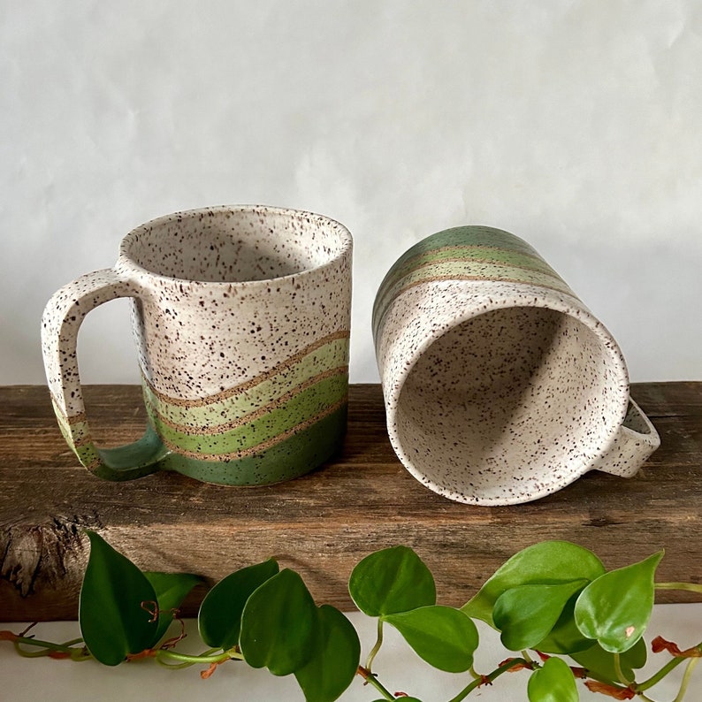 May include: Two ceramic mugs with a speckled white glaze and green and brown stripes. The mugs are sitting on a wooden surface with green plants in the foreground.