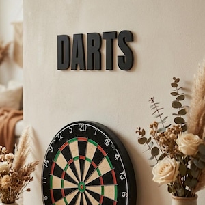 Könnte beinhalten: Eine schwarze Dartscheibe mit grün-roter Mitte ist an einer beigen Wand befestigt. Das Wort "DARTS" ist in schwarzen Blockbuchstaben über der Dartscheibe zu sehen. Dekorative Vasen mit Blumen befinden sich im Vordergrund.