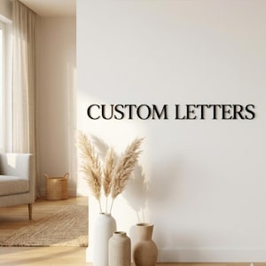 Könnte beinhalten: Schwarze, individuelle Buchstaben an einer weißen Wand. Der Text lautet „CUSTOM LETTERS“. Die Buchstaben sind in einer serifenlosen Schriftart gehalten. Die Wand ist mit getrocknetem Pampasgras in weißen und beigen Vasen und einem geflochtenen Korb dekoriert.