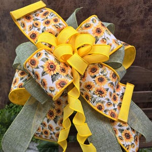 May include: A decorative bow featuring a sunflower pattern on a white background, accented with yellow and green ribbons. The bow has multiple loops and streamers, creating a full, dimensional look. The sunflower design includes yellow petals and brown centers.