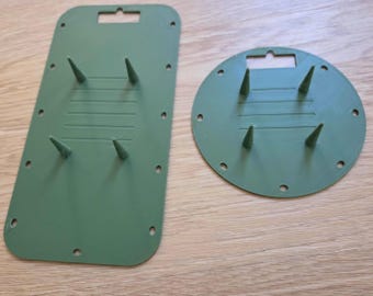 May include: Two olive green plastic floral foam holders, one rectangular and one circular. Both feature four pointed spikes for securing foam, multiple small holes along the edges for attachment, and a rectangular hanging slot at the top.