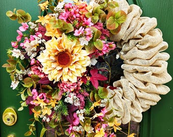 May include: A fall-themed wreath with a burlap base and a variety of flowers, including sunflowers, pink hydrangeas, and burgundy leaves.