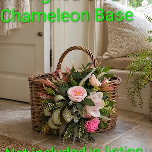 K&ouml;nnte beinhalten: Ein geflochtener brauner Weidenkorb mit einem dekorativen Arrangement aus rosa Rosen, wei&szlig;en Blumen, gr&uuml;nem Blattwerk und einem strukturierten olivgr&uuml;nen Band. Der Text "Designed on a Chameleon Base" und "Not included in listing" ist in leuchtendem Gr&uuml;n &uuml;berlagert.