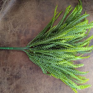 May include: A bundle of artificial fern leaves in varying shades of green. The leaves are long and slender, with a delicate, textured appearance. The stems are a darker green, and the overall effect is a natural, decorative element.