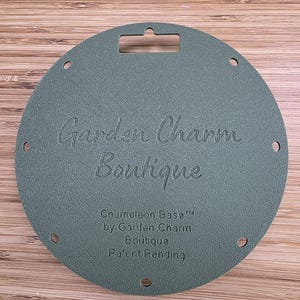 May include: A circular, sage green textured base with a rectangular hanging slot at the top and eight small holes around the perimeter. The text Garden Charm Boutique, Chameleon Base, and Patent Pending is embossed on the front surface.