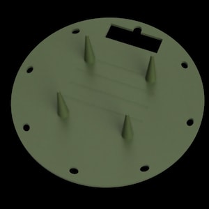 May include: A circular olive green plate featuring four conical spikes protruding from the center, a rectangular cutout near the top edge, and a series of small circular holes spaced evenly around the outer perimeter of the disc.
