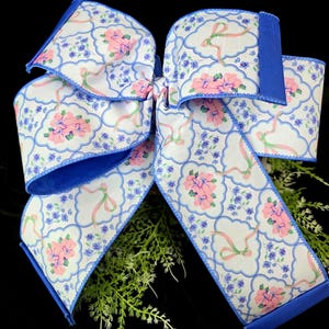 May include: A large decorative bow with a floral pattern. The bow features a white background with pink flowers, blue accents, and a blue border. The bow is made of fabric and is suitable for various decorative purposes.