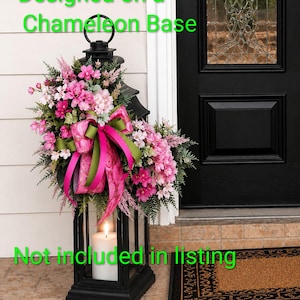 K&ouml;nnte beinhalten: Eine schwarze Metalllaterne, verziert mit einem Blumenarrangement aus rosa und wei&szlig;en Bl&uuml;ten, gr&uuml;nem Blattwerk und einem gemusterten rosa-gr&uuml;nen Band. Eine wei&szlig;e Stumpenkerze steht darin. Text: Designed on a Chameleon Base. Not included in listing.