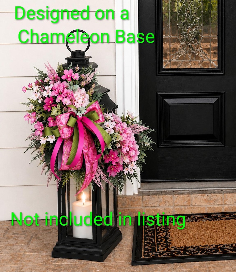K&ouml;nnte beinhalten: Eine schwarze Metalllaterne, verziert mit einem Blumenarrangement aus rosa und wei&szlig;en Bl&uuml;ten, gr&uuml;nem Blattwerk und einem gemusterten rosa-gr&uuml;nen Band. Eine wei&szlig;e Stumpenkerze steht darin. Text: Designed on a Chameleon Base. Not included in listing.