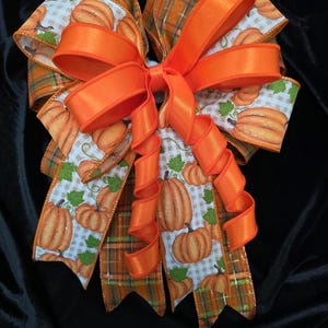 May include: A decorative bow featuring a combination of orange satin ribbon and a patterned ribbon with orange pumpkins, green leaves, and a light brown checkered background. The bow has multiple loops and long, curled ribbon tails.