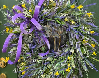 May include: A decorative wreath featuring lavender, yellow, and white flowers with green foliage. A large purple velvet ribbon is tied in a bow at the top. The wreath is hanging on a green door.