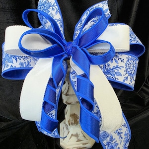 May include: A decorative bow featuring white and blue floral patterned ribbon, accented with solid royal blue ribbon. The bow is centered on a distressed white decorative object, creating a layered effect. The bow's design is suitable for various decorative applications.
