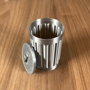 May include: A small, silver-colored trash can with a matching lid. The can has vertical, rounded grooves around its exterior. The lid is round with a star-shaped handle. The items are made of a metallic material and sit on a wooden surface.