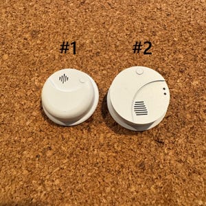 Puede incluir: Dos detectores de humo blancos sobre un fondo marrón. Uno, etiquetado como "#1", tiene una rejilla de altavoz. El otro, etiquetado como "#2", presenta ranuras horizontales y dos pequeños puntos.