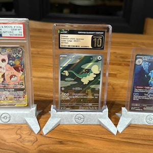 May include: Three graded Pokémon trading cards in protective cases, displayed on gray stands. The cards feature colorful illustrations of various Pokémon characters. The stands have a Pokémon logo.