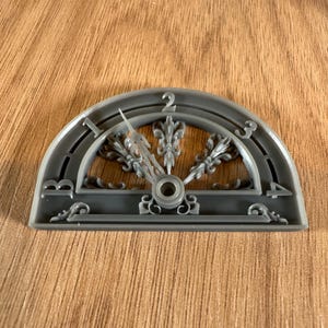 May include: A gray, semi-circular decorative clock face with the numbers 1, 2, 3, and 4. The clock face has an ornate design with a central dial and a decorative needle. The background is a light brown wood grain.