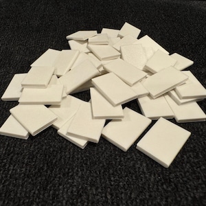 May include: A collection of small, rectangular, off-white tiles scattered on a dark, textured surface. The tiles appear to be made of a solid material, possibly ceramic or a similar substance, and are uniform in shape and size.