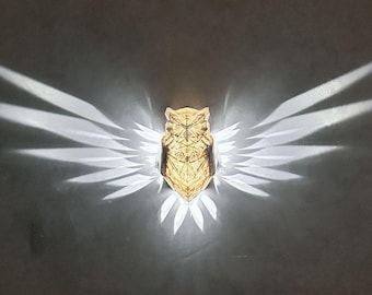 May include: A gold owl-shaped wall lamp with geometric patterns casts a white light onto a wall, creating a shadow of wings.