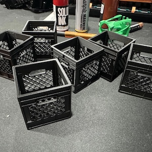 May include: Six black plastic mini crates, stacked and arranged in a staggered pattern on a gray surface.