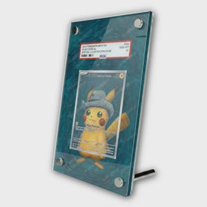 May include: A framed Pokémon card featuring Pikachu wearing a hat. The card is encased in a clear acrylic display with a teal background. Text on the card reads "Pika-Portrait". The frame has silver hardware and a stand.