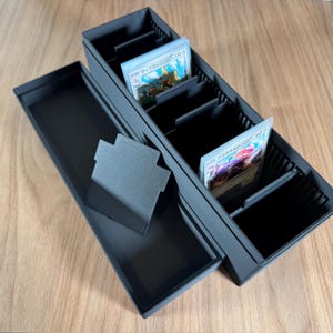 May include: A black, multi-compartment card storage box with a lid. The box is open, revealing several sections holding trading cards. The cards have Japanese text and colorful illustrations. A separate, small, black card holder is also present.