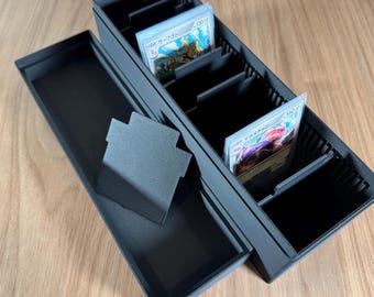 Puede incluir: Una caja negra de almacenamiento de cartas con múltiples compartimentos y una tapa. La caja está abierta, mostrando cartas coleccionables con texto e ilustraciones japonesas. También hay un pequeño soporte negro para tarjetas separado.
